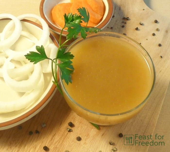 Beef broth in a small bowl next to sliced onions and carrots and garnished with parsley