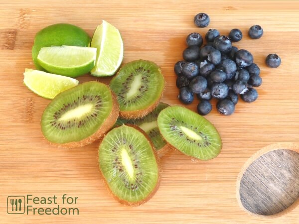 Sliced limes and kiwis and a small pile of blueberries on a bamboo cutting board