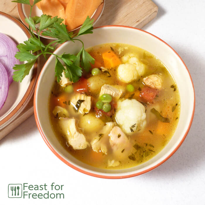 Chicken soup in a bowl next to fresh carrots and onions, and garnished with parsley