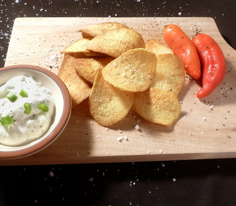 Potato chips on a wooden board, sprinkled with coarse salt and freshly ground pepper, next to a bowl of chip dip